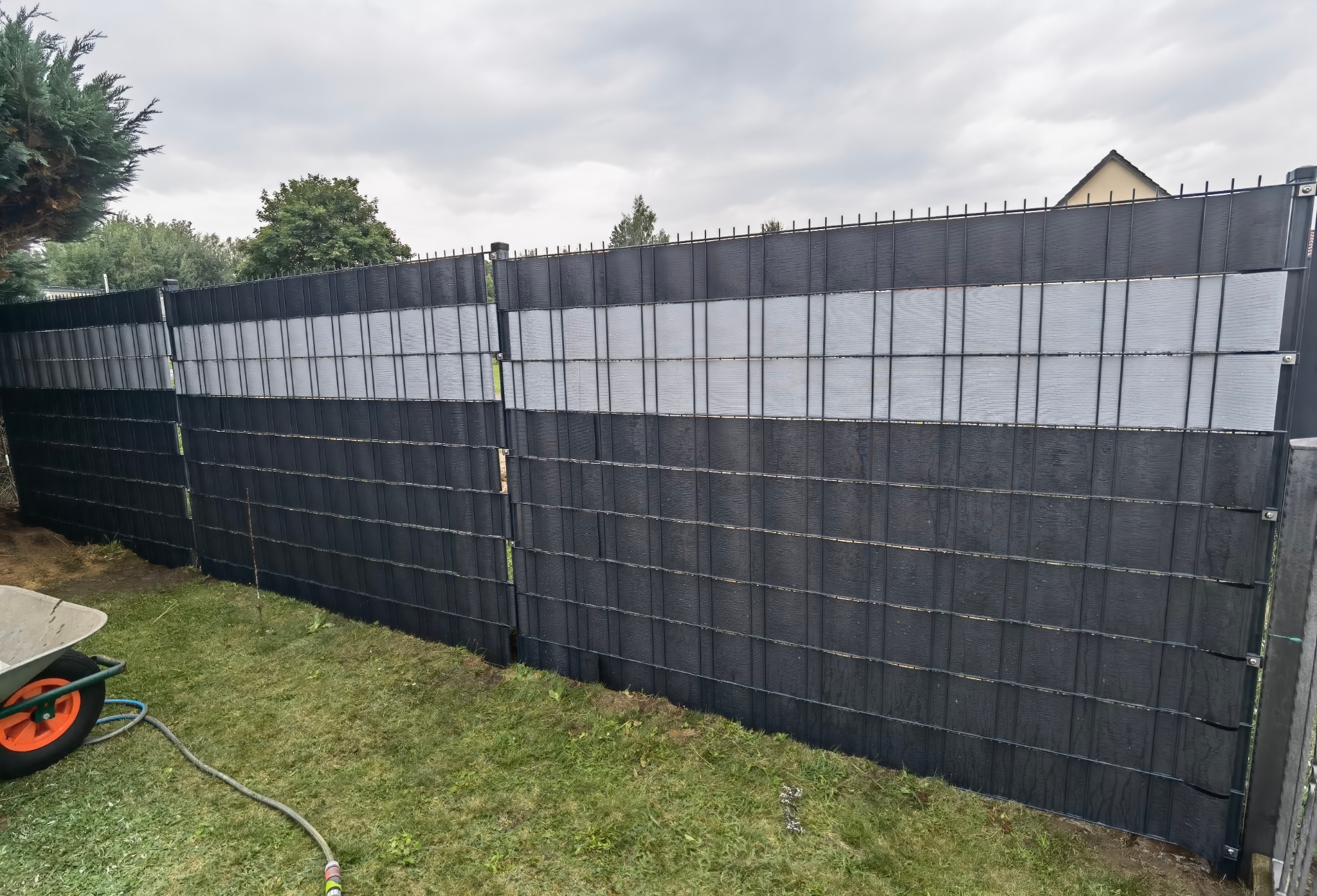 Nowoczesne ogrodzenie panelowe z poziomymi pasami w kolorze czarnym i szarym, widok z zewnątrz na tle zielonego trawnika i pochmurnego nieba, z taczką ogrodową w lewym dolnym rogu.