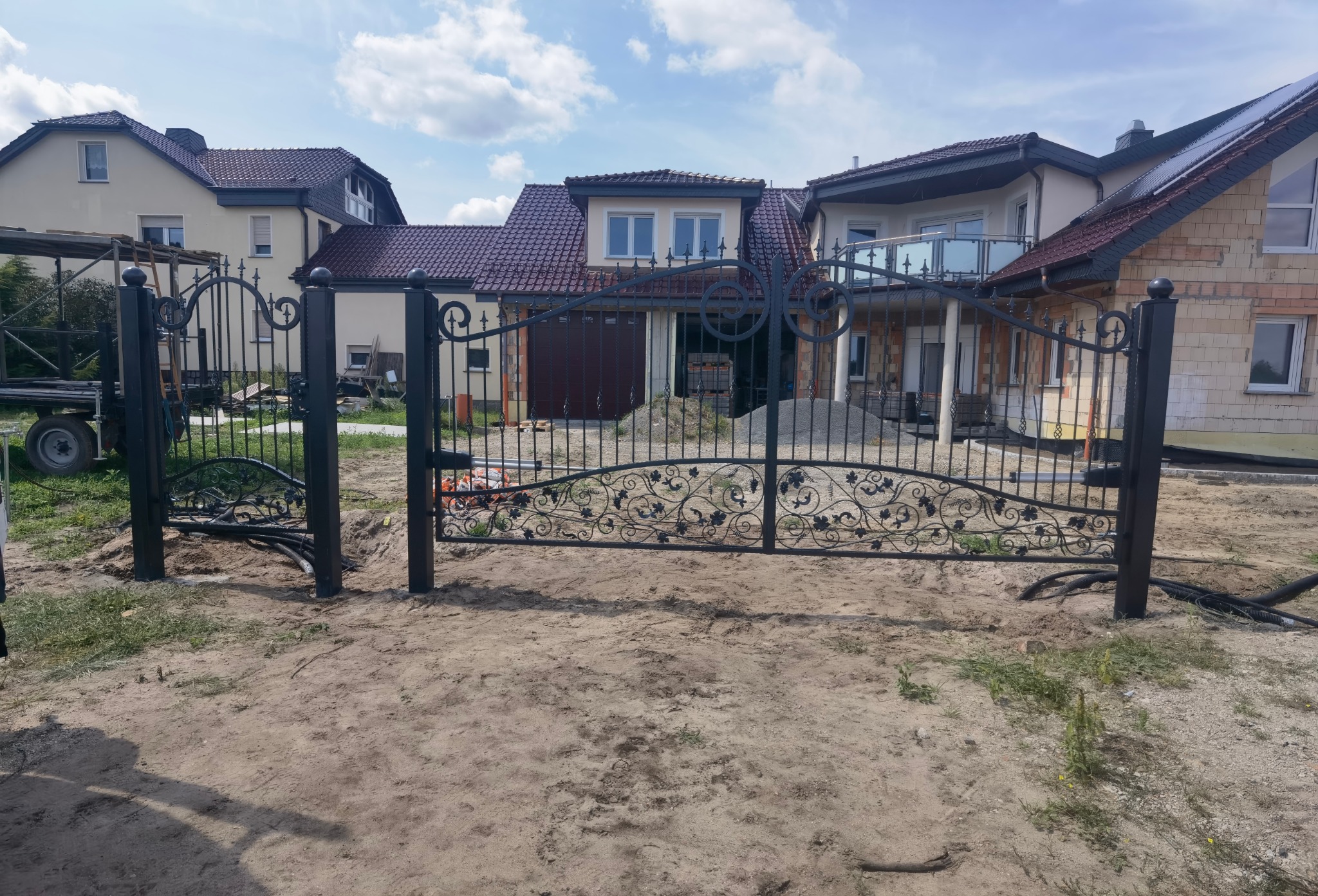 Kuta brama wjazdowa z ozdobnymi elementami roślinnymi, świeżo zamontowana na piaszczystym terenie budowy, widoczne kable elektryczne do automatyki, dom w tle.