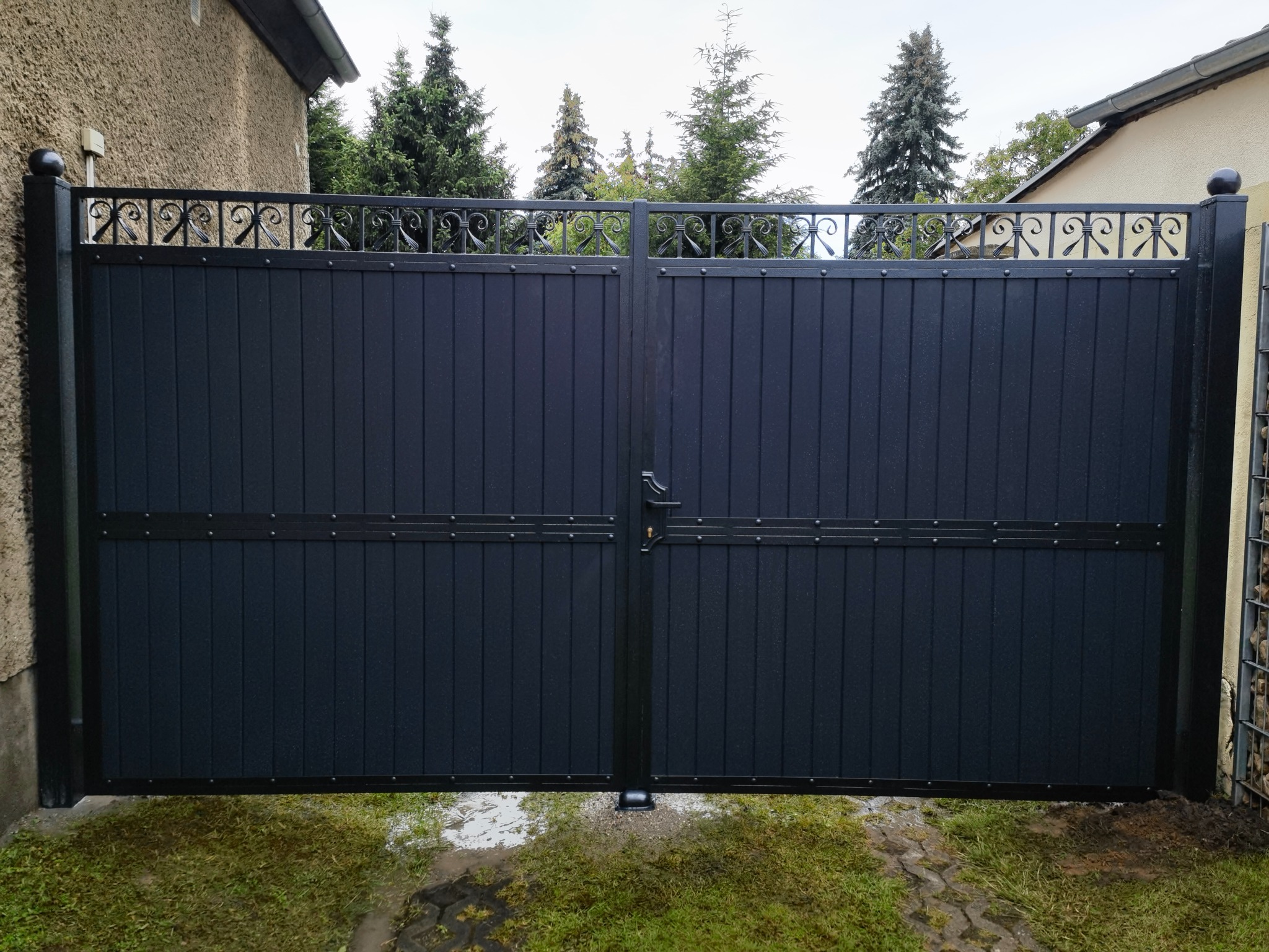 Dwuskrzydłowa brama wjazdowa, ciemna, z metalowymi panelami pionowymi i ozdobnym, kutym wzorem u góry, widok z zewnątrz na tle zielonych drzew.