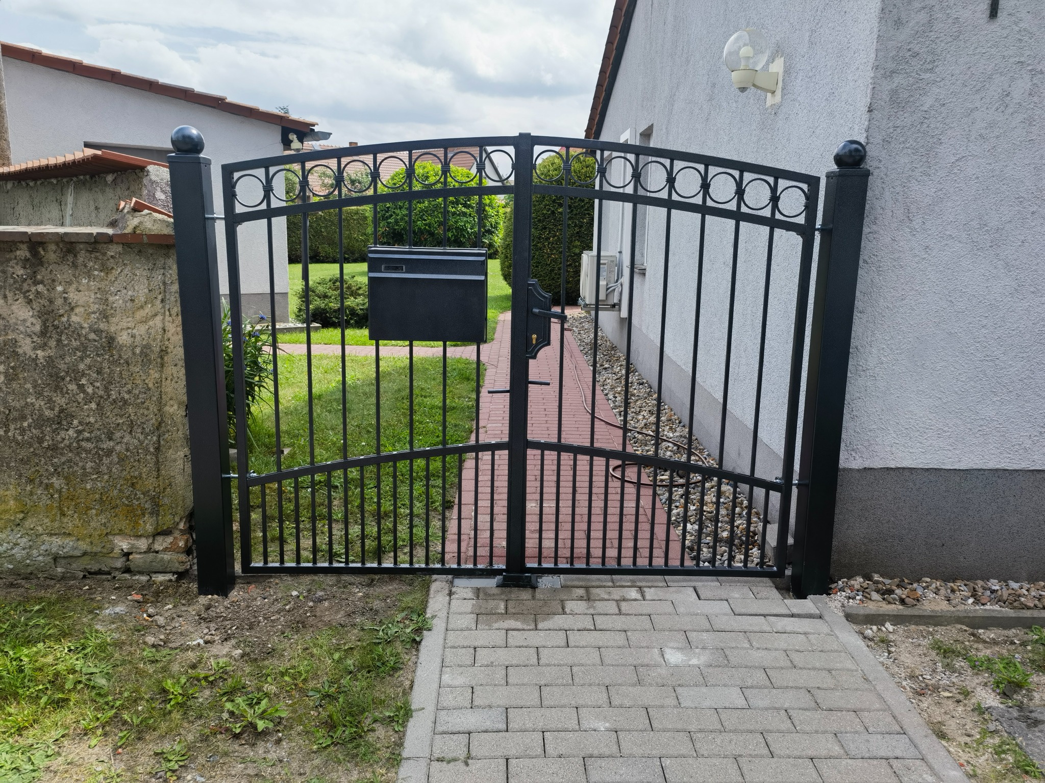 Czarna, metalowa brama dwuskrzydłowa z okrągłym wzorem, prowadząca do domu z ceglaną ścieżką i skrzynką na listy.