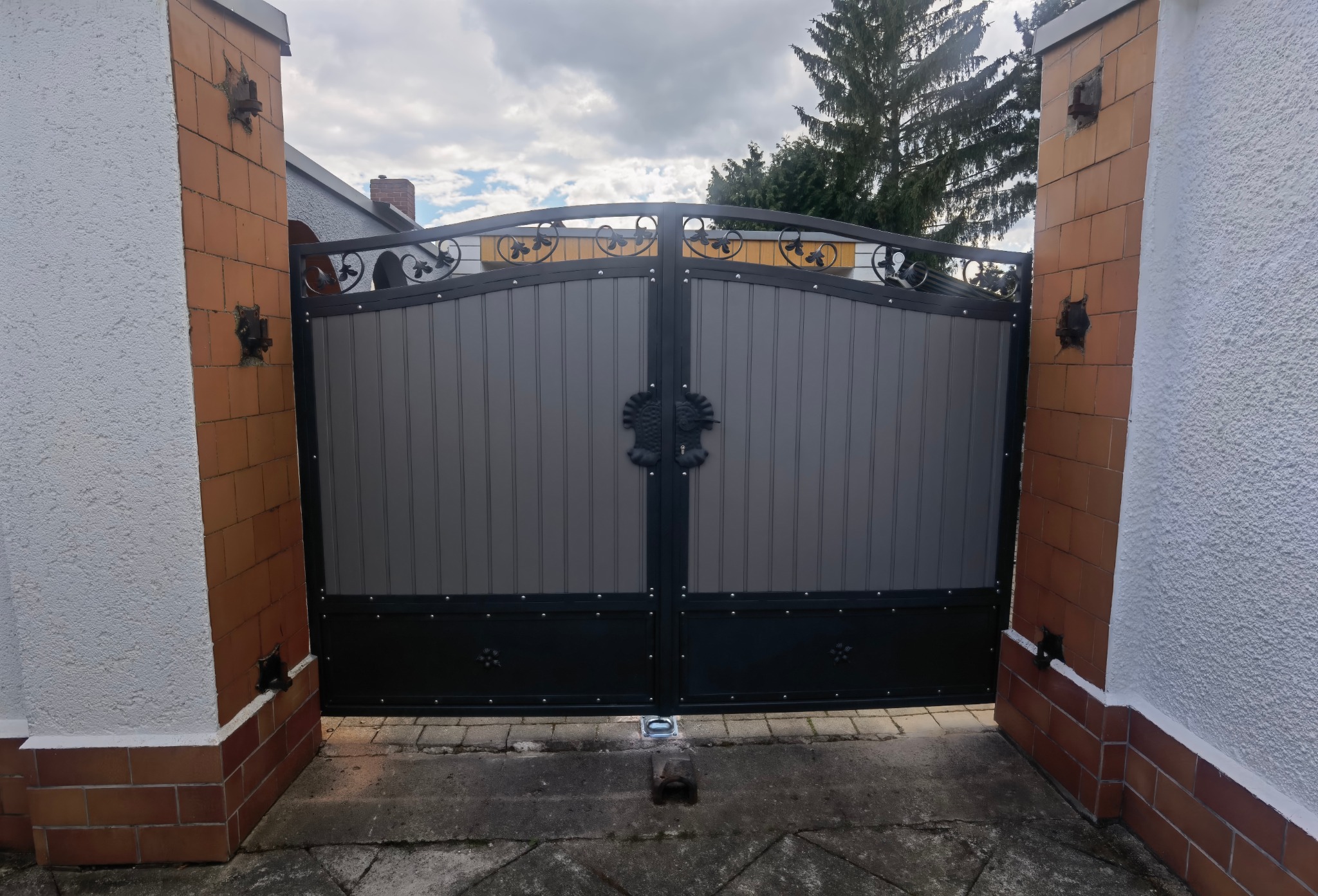 Dwuskrzydłowa brama wjazdowa z szarymi panelami i ozdobnymi elementami metalowymi, mocowana do murowanych słupków z cegieł.