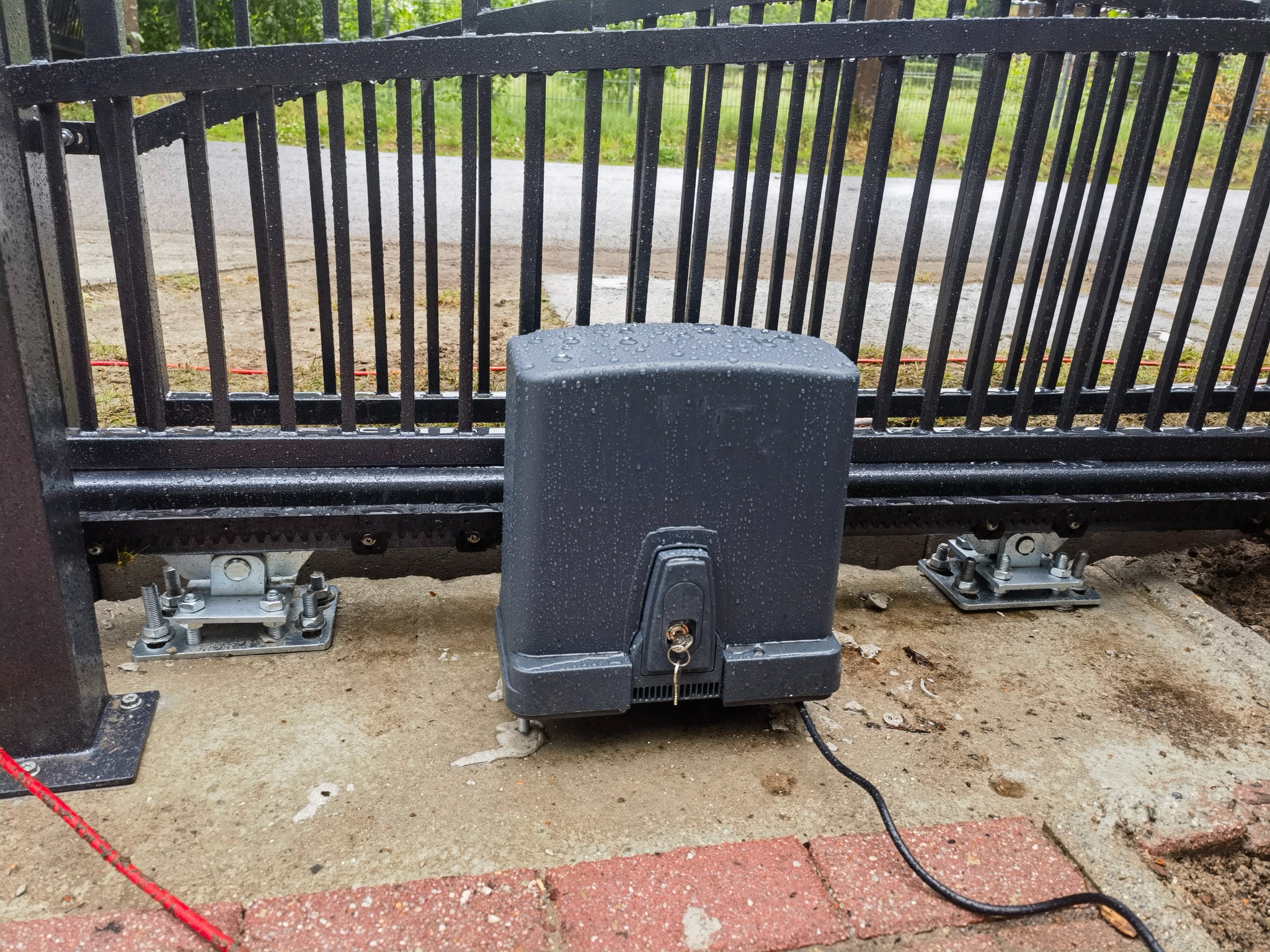 Czarny automat do bramy przesuwnej, widoczne krople deszczu, klucz w zamku, szyna bramy i kostka brukowa.