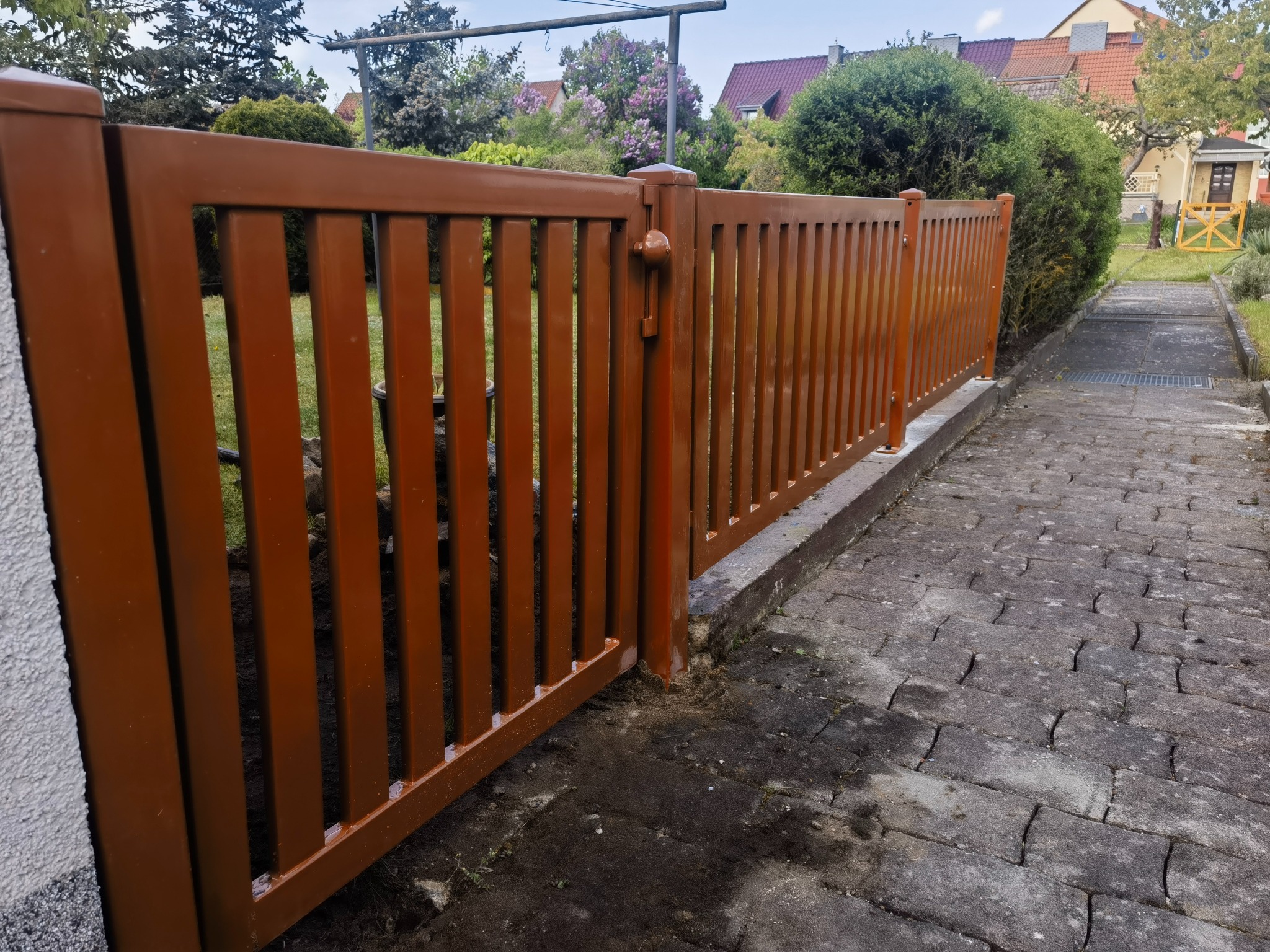 Otwarta brązowa furtka z pionowych metalowych prętów, widoczny mechanizm zamknięcia, w tle ogród z kwitnącymi bzami i domy.