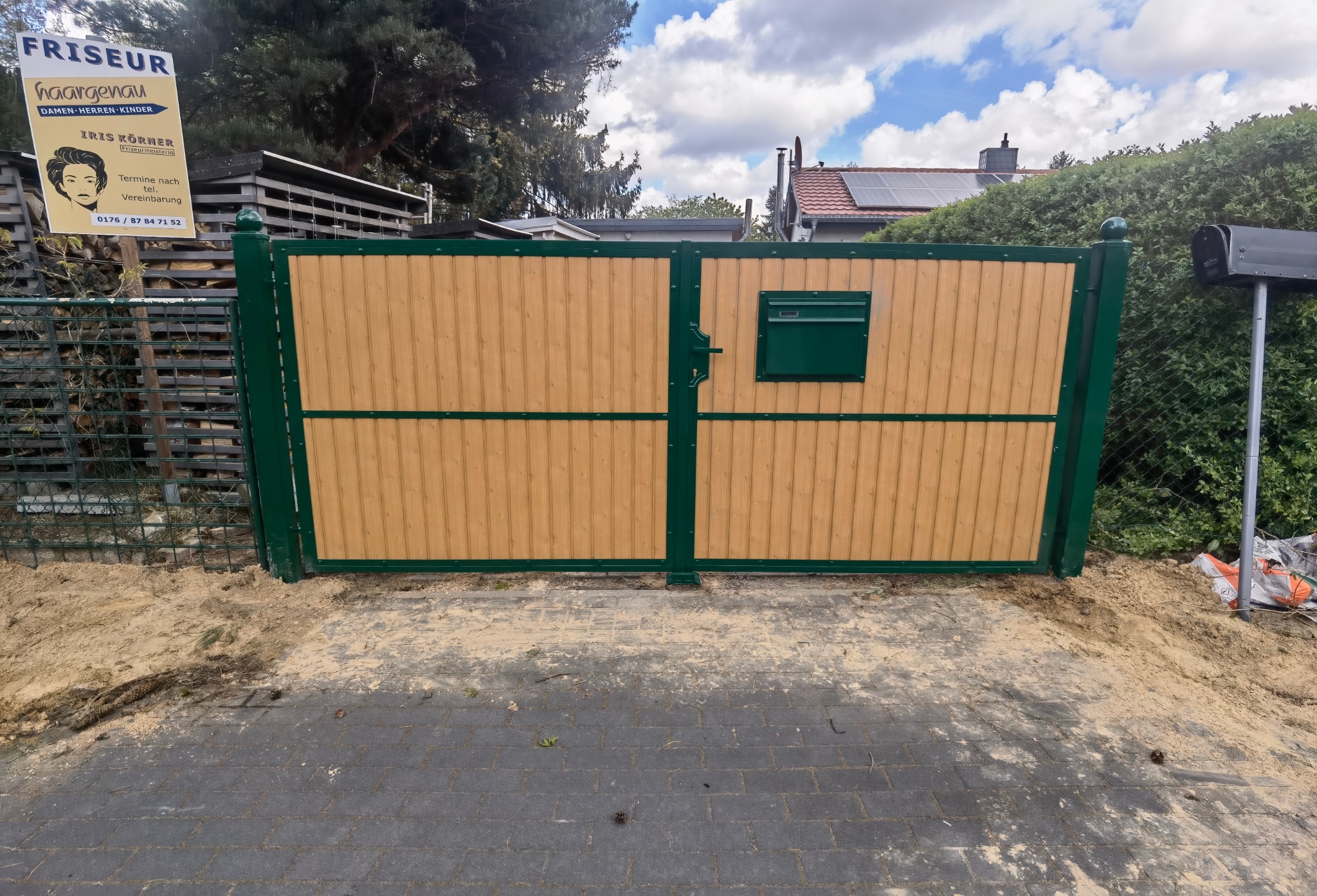 Zamknięta brama dwuskrzydłowa z drewna i metalu w kolorze zielonym, z zamontowaną skrzynką pocztową, widoczna z zewnątrz na tle zieleni i niebieskiego nieba, świeżo po montażu - widać piasek.