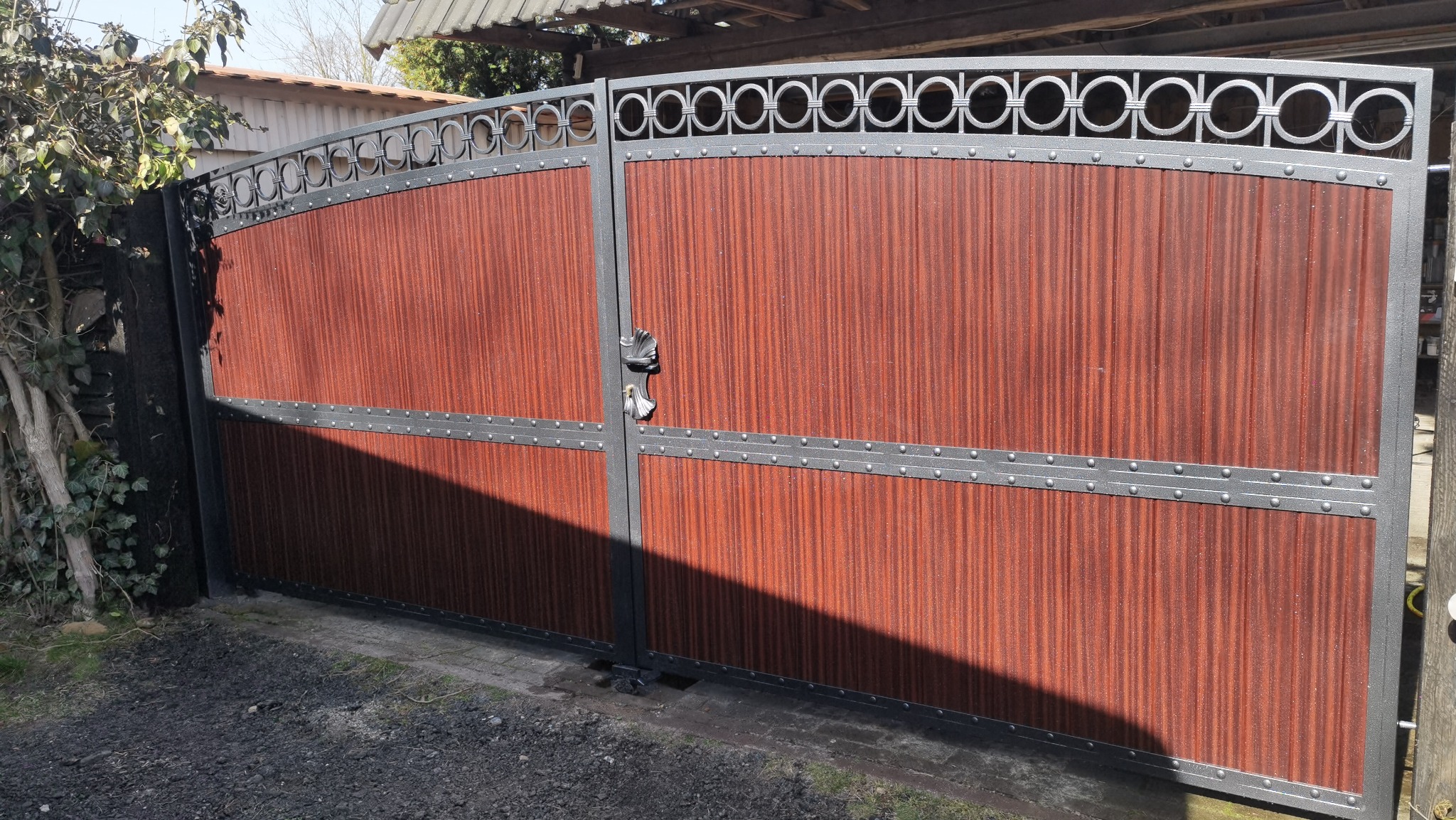 Dwuskrzydłowa brama wjazdowa z metalową ramą i wypełnieniem z paneli imitujących drewno, ozdobiona motywem kółek w górnej części, z widocznym zamkiem i cieniem na jednym ze skrzydeł.