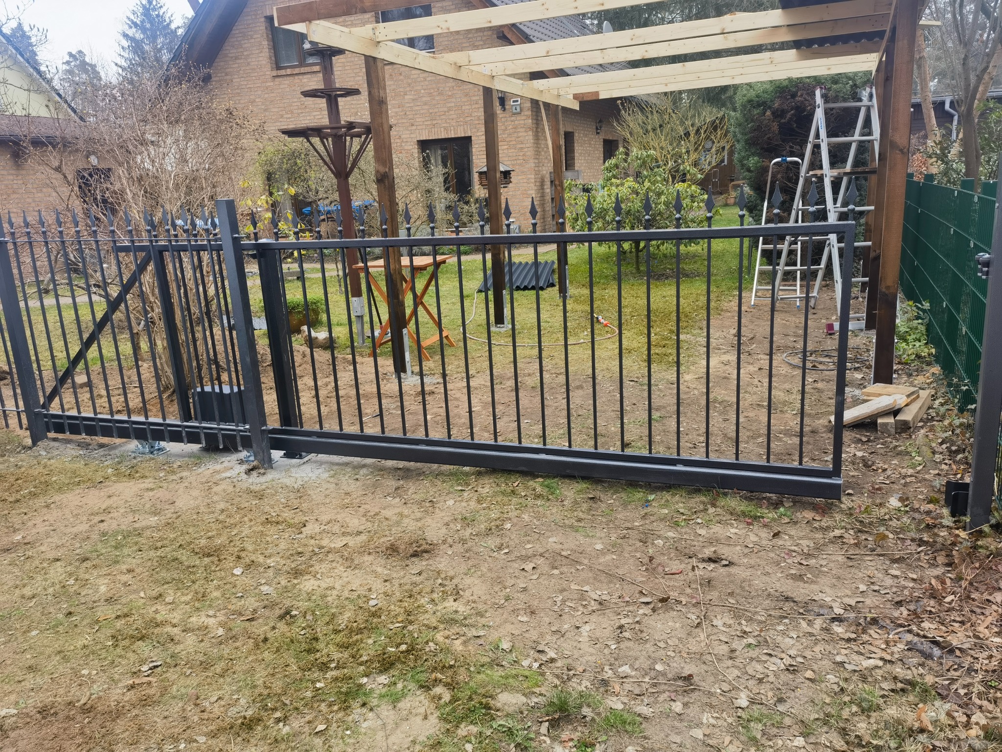 Czarna, metalowa brama przesuwna w trakcie montażu, widoczne elementy mocujące do podłoża, w tle konstrukcja drewnianej pergoli i dom z cegły.