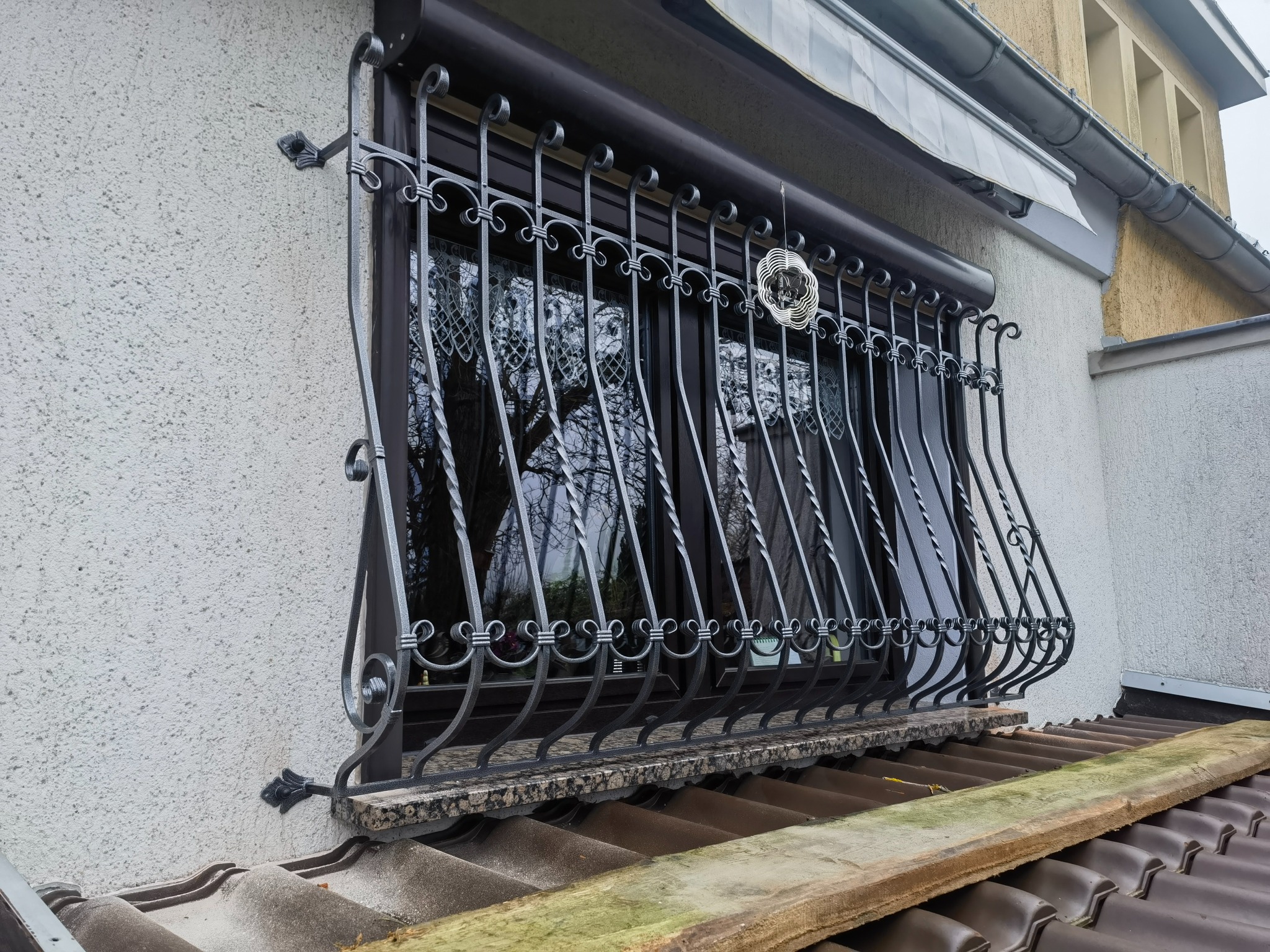 Okno zabezpieczone ozdobną, metalową kratą z dekoracyjnymi elementami, widoczne z zewnątrz, z drewnianą deską opartą o dachówkę.