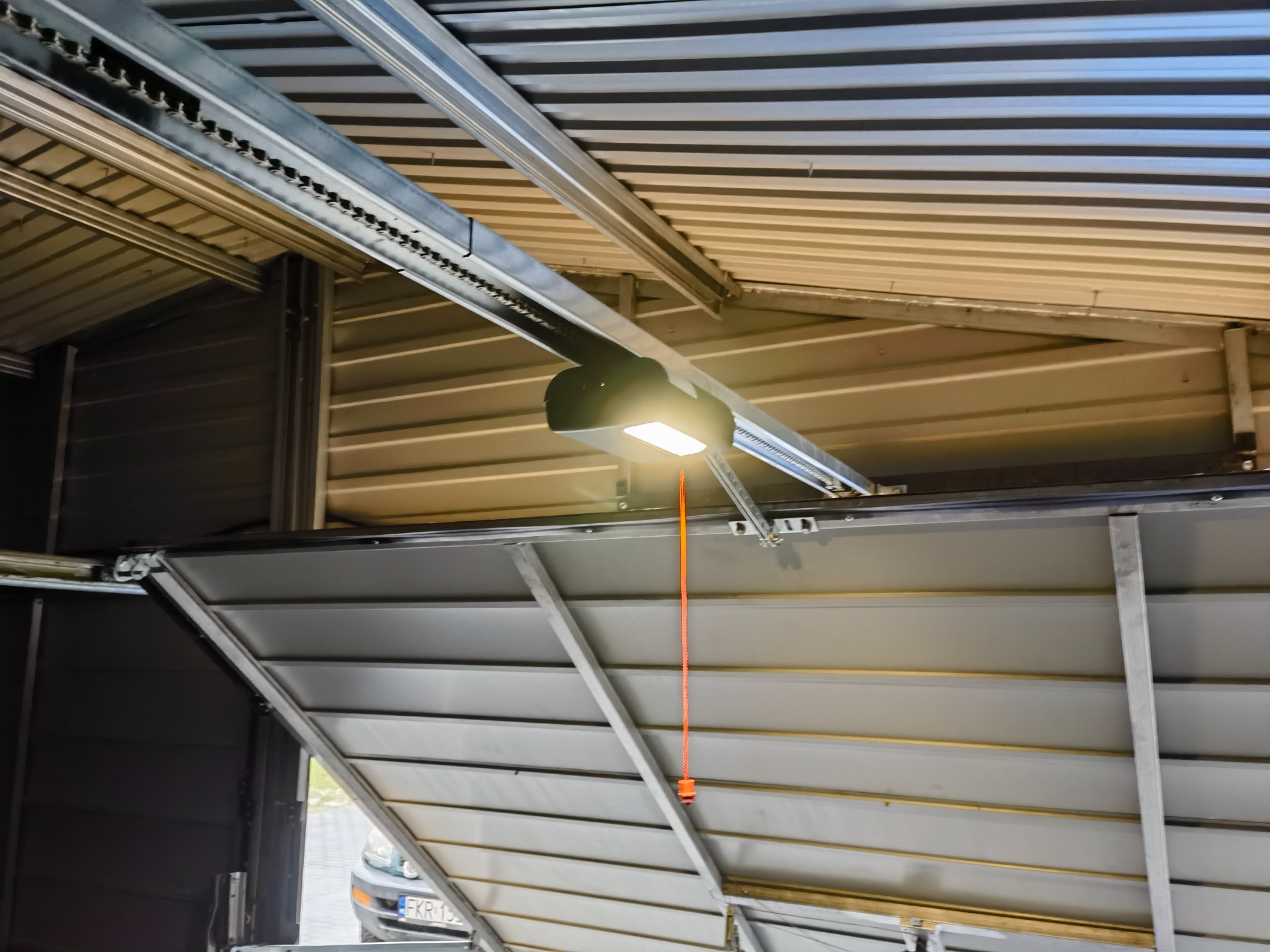 Wnętrze garażu z widocznym mechanizmem napędu bramy garażowej, łańcuch, silnik z oświetleniem i częściowo otwarta brama segmentowa.