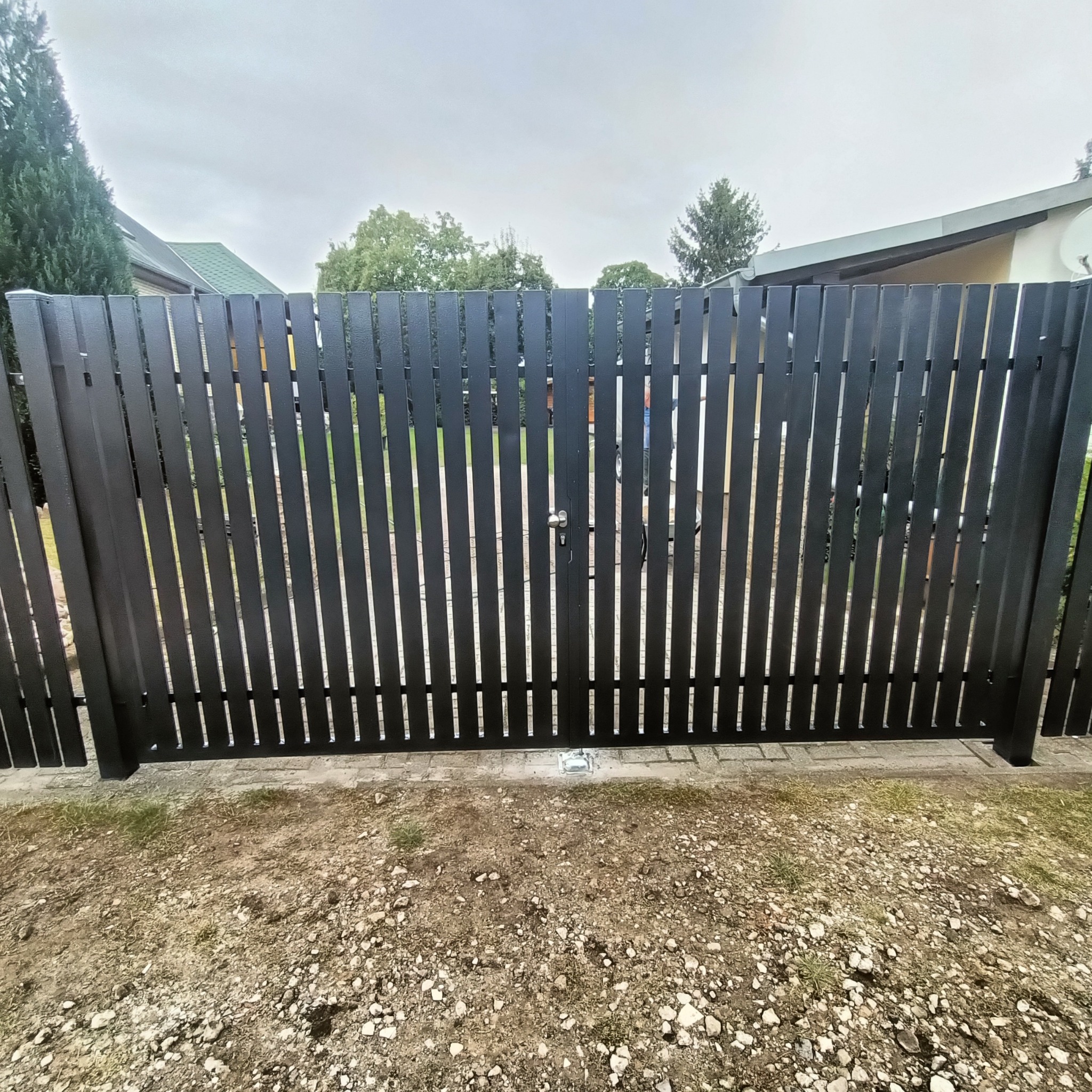Nowoczesna, czarna brama z pionowych profili, zabezpieczona zamkiem, na tle domu i drzew pod pochmurnym niebem.