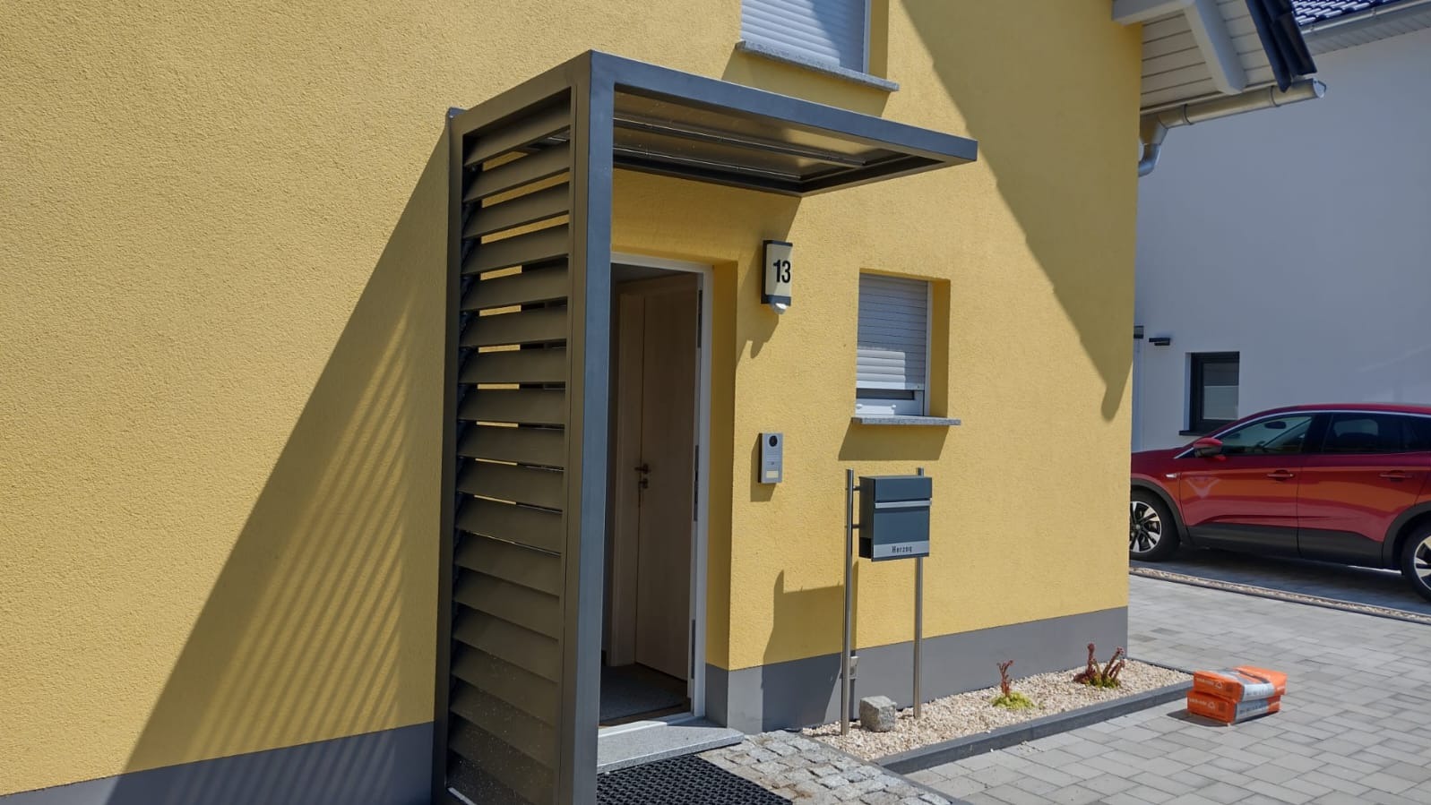 Nowoczesne zadaszenie nad wejściem do domu z lamelami, skrzynka na listy i elewacja w kolorze żółtym, widok na podjazd i auto.
