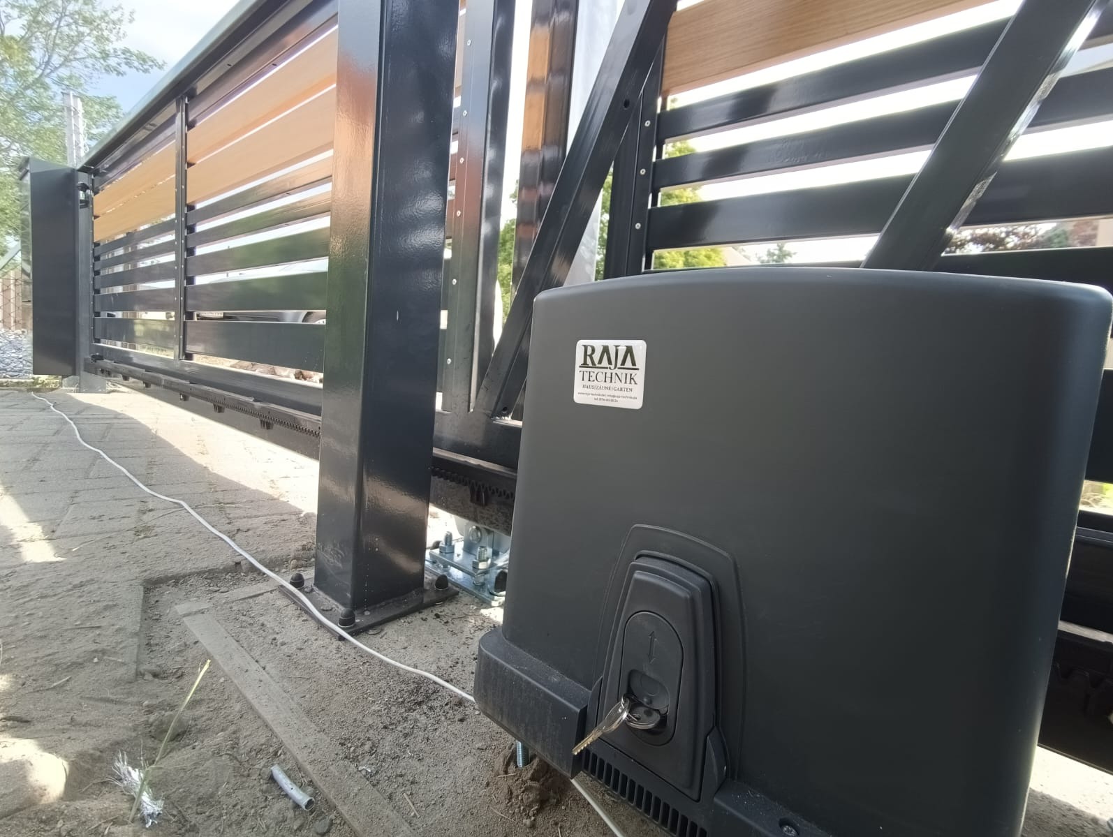 Zasuwna brama z drewnianymi elementami i czarną metalową ramą, widoczny automat z kluczykiem w zamku, kabel zasilający i podłoże z kostki brukowej.