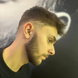 Męska fryzura z cieniowaniem i delikatnym wzorkiem na skroni, widok z profilu na tle ściany z abstrakcyjnym wzorem.