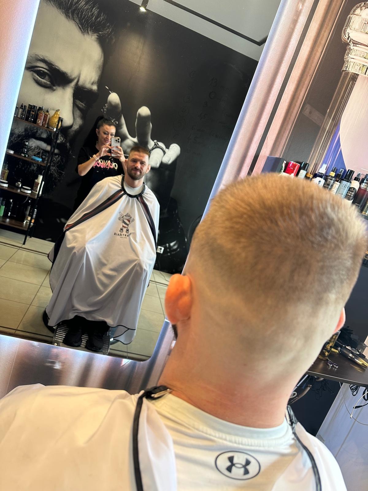 Krótko ostrzyżony mężczyzna w pelerynie fryzjerskiej, widziany z tyłu głowy, odbity w lustrze, gdzie fryzjerka robi mu zdjęcie telefonem na tle ściany z wizerunkiem brodatego mężczyzny i półkami...