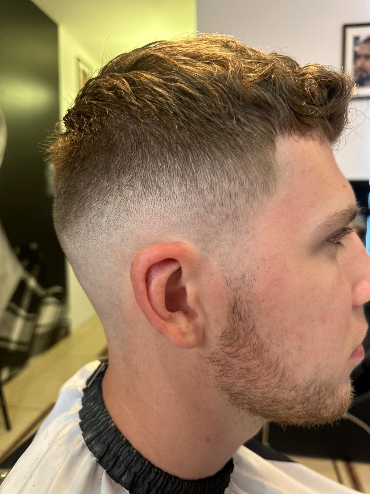 Męska fryzura z wycieniowanymi bokami i krótko przystrzyżoną brodą, widok z profilu, podczas zabiegu w salonie.