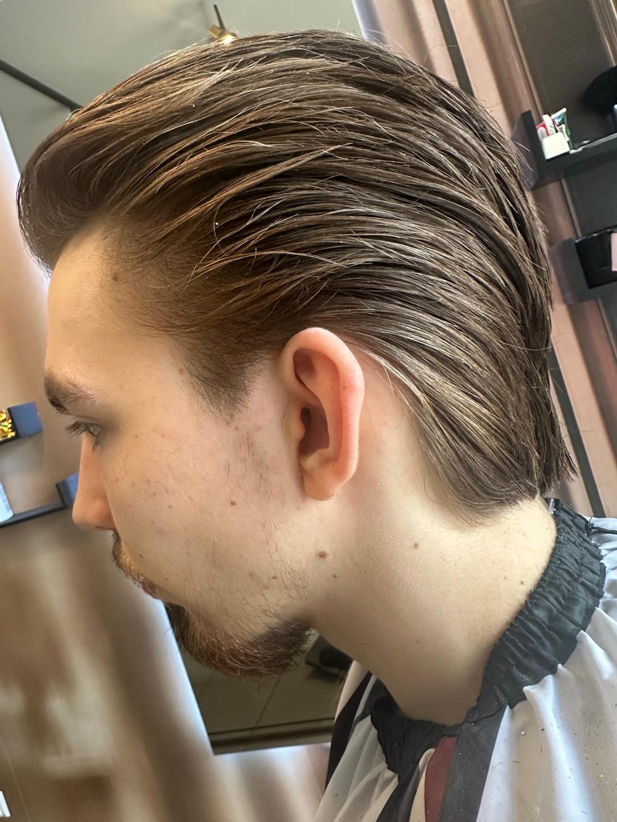 Męska fryzura z zaczesanymi do tyłu, brązowymi włosami, widok z profilu na tle salonu.