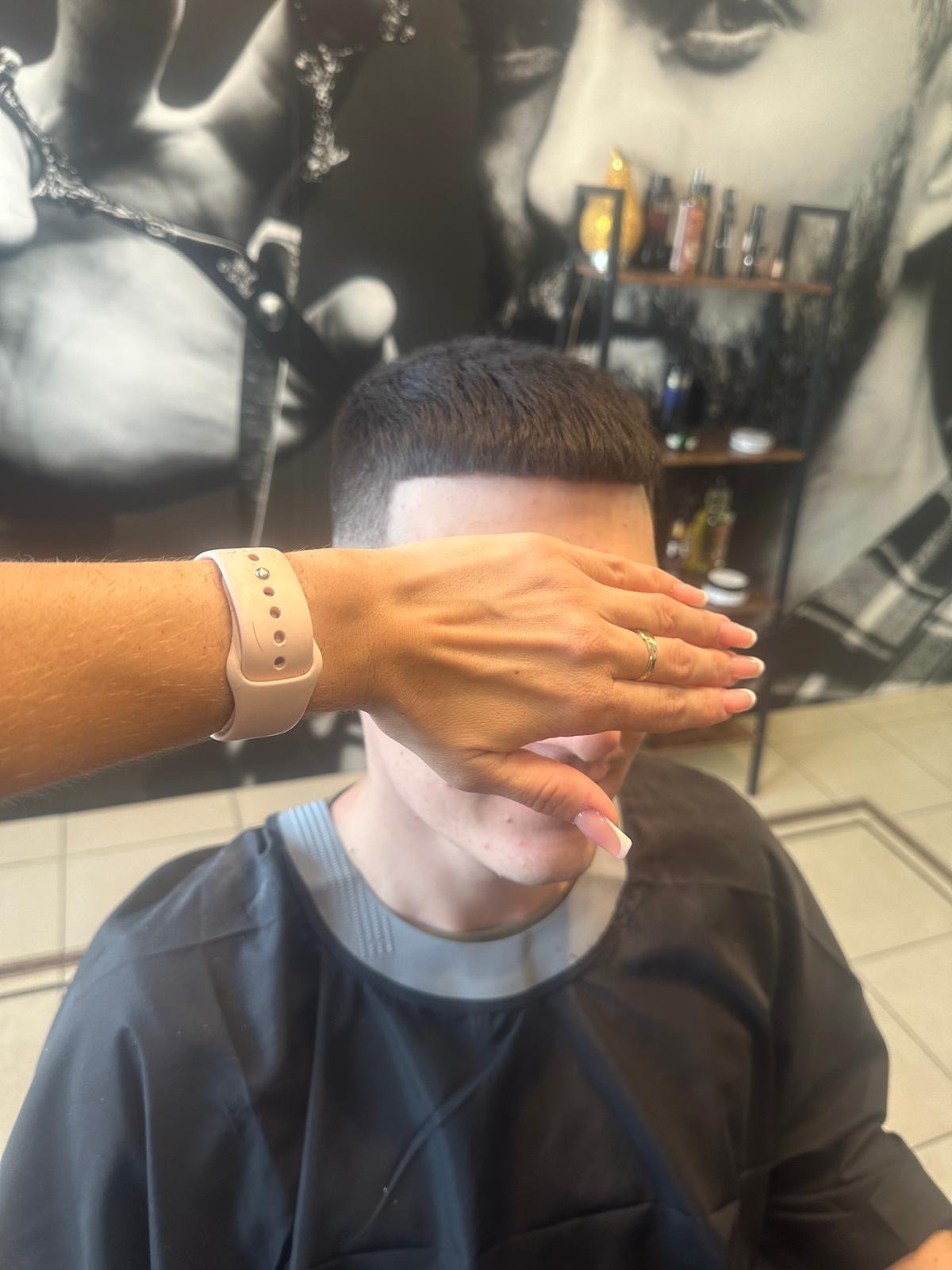 Mężczyzna z krótką fryzurą zasłaniający twarz dłonią stylistki z beżowym smartwatchem i złotym pierścionkiem, w tle półka z kosmetykami i czarno-biała grafika.