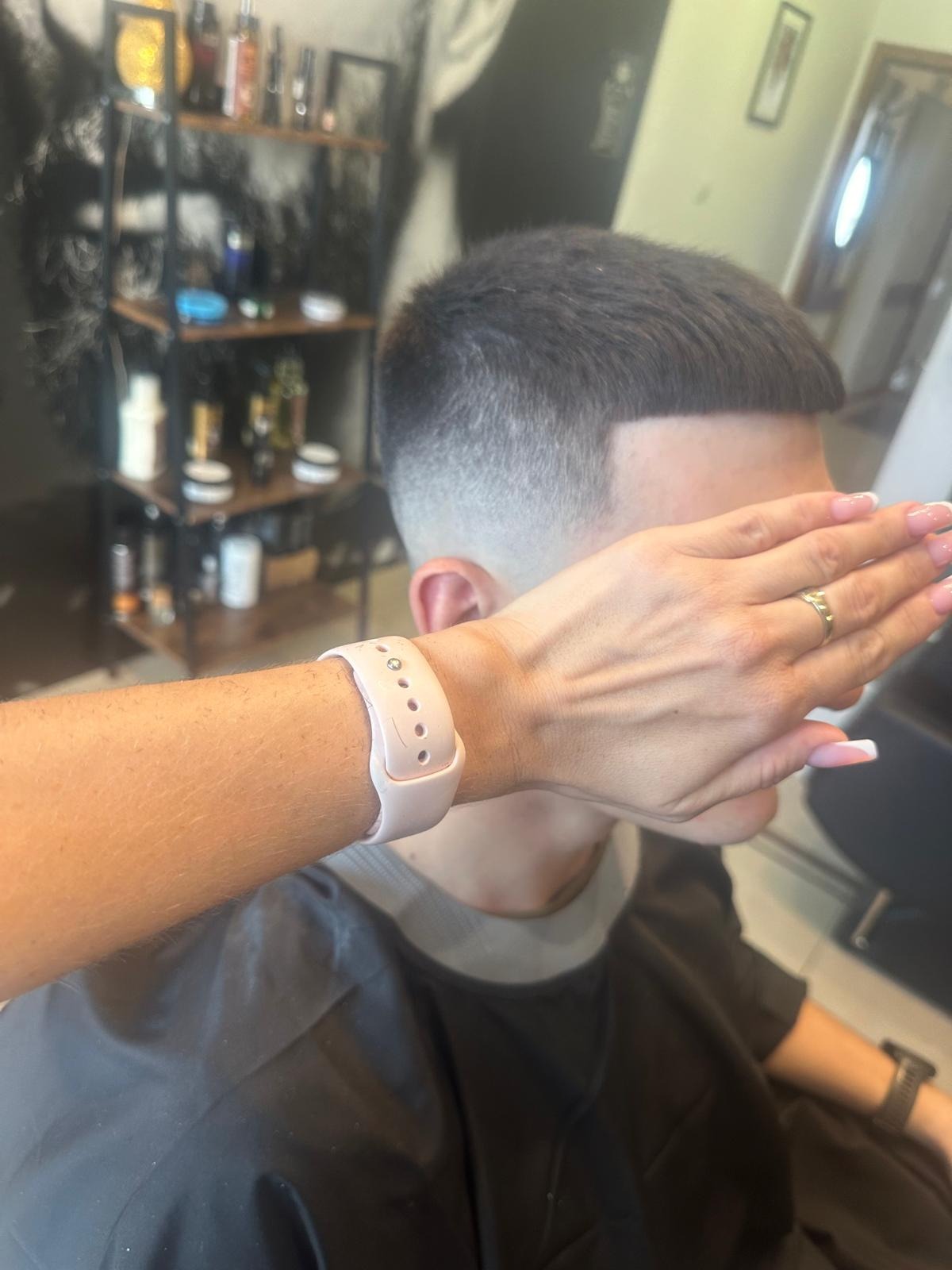 Męskie strzyżenie z cieniowaniem, ręka z różowym smartwatchem zasłania twarz klienta, widoczny fragment dłoni z manicure hybrydowym i złotym pierścionkiem, w tle półki z kosmetykami.