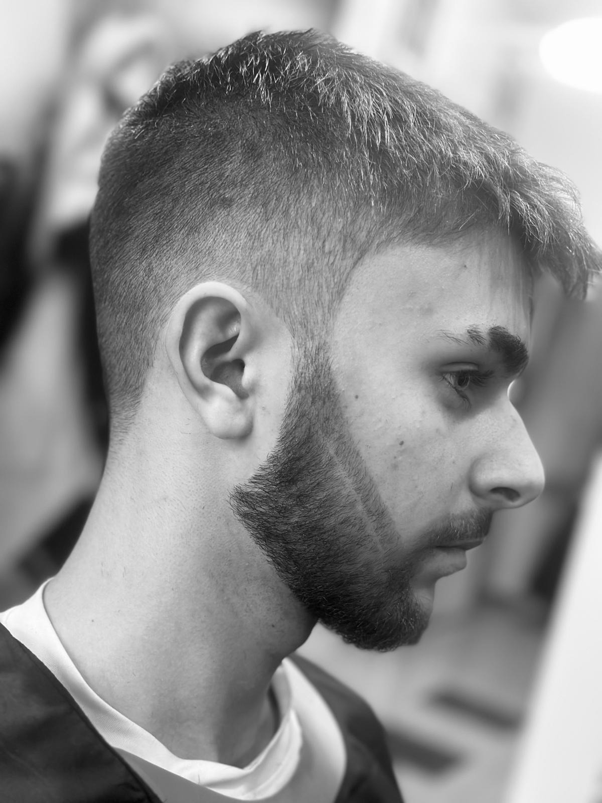Profil młodego mężczyzny z precyzyjnie wystylizowaną brodą i krótką fryzurą w odcieniach szarości, ukazujący detale strzyżenia i modelowania zarostu.