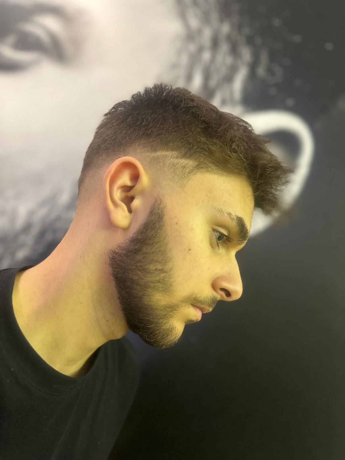 Męska fryzura z cieniowaniem i delikatnym wzorkiem na skroni, widok z profilu na tle ściany z abstrakcyjnym wzorem.