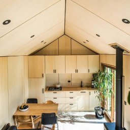 WoodWorld - Świat Drewna - Wnętrze domu z drewnianą konstrukcją szkieletową, widok na kuchnię z drewnianymi szafkami, stół z krzesłami i piec wolnostojący. Charakterystyczny skośny sufit z wbudowanymi punktami świetlnymi.
