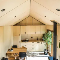 Wnętrze domu z drewnianą konstrukcją szkieletową, widok na kuchnię z drewnianymi szafkami, stół z krzesłami i piec wolnostojący. Charakterystyczny skośny sufit z wbudowanymi punktami świetlnymi.