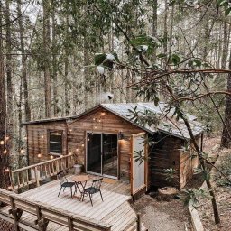 WoodWorld - Świat Drewna - Drewniany domek w lesie z tarasem, oświetlony girlandami, z widocznym dymem z komina i śniegiem na gałęziach, widziany z góry.