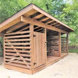 Drewniana konstrukcja z zamykanymi drzwiami, prawdopodobnie skład na drewno, z poziomymi listwami tworzącymi ażurową ścianę, na tle zieleni.