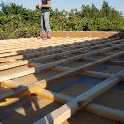 Ludwik  Grudzień - Konstrukcja drewniana szkieletu budynku w trakcie budowy, widoczne belki i izolacja, na górze stoi osoba w jeansach i czerwonych butach.