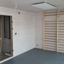 WoodWorld - Świat Drewna - Wnętrze pomieszczenia wykończonego boazerią w jasnym odcieniu, z zamontowanymi drabinkami gimnastycznymi, przeszklonymi drzwiami i widocznymi elementami instalacji elektrycznej w trakcie montażu.