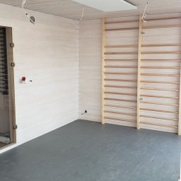 WoodWorld - Świat Drewna - Wnętrze pomieszczenia wykończonego drewnem z drabinkami gimnastycznymi, szarą podłogą i panelami sufitowymi w trakcie montażu oświetlenia.