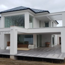 WoodWorld - Świat Drewna - Nowoczesny, dwukondygnacyjny dom z płaskim dachem i dużymi oknami, zadaszony taras z szarych desek kompozytowych, widoczne prace wykończeniowe i materiały budowlane na zewnątrz.