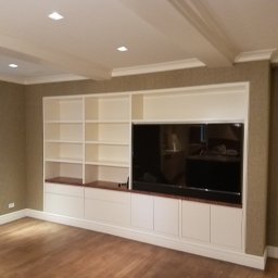 Ludwik  Grudzień - Biała zabudowa meblowa z półkami i szafkami w salonie z telewizorem, drewniana podłoga, dekoracyjne listwy sufitowe, ściany w odcieniach beżu, widoczna część drabiny malarskiej.