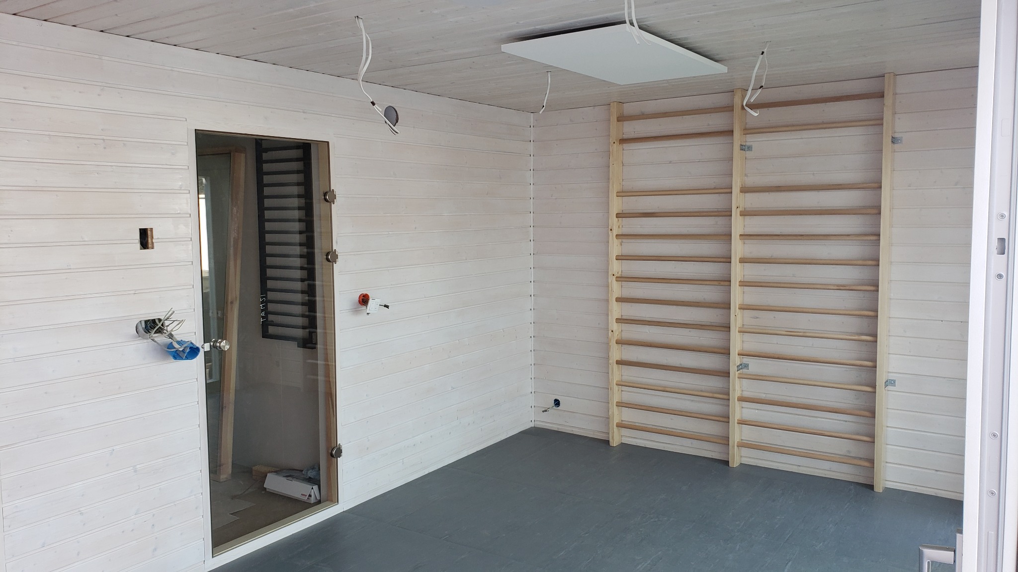 Wnętrze pomieszczenia wykończonego boazerią w jasnym odcieniu, z zamontowanymi drabinkami gimnastycznymi, przeszklonymi drzwiami i widocznymi elementami instalacji elektrycznej w trakcie montażu.