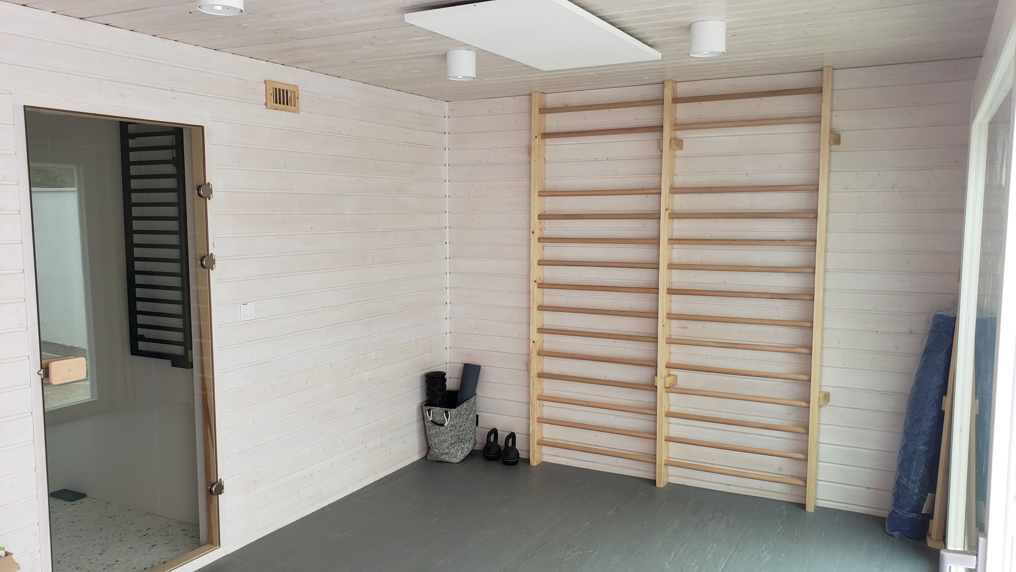 Jasne wnętrze sauny z drewnianymi panelami, szklanymi drzwiami, drabinką gimnastyczną i koszem z akcesoriami do ćwiczeń.