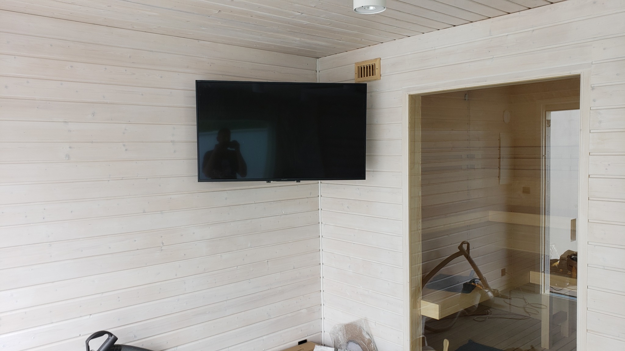 Wnętrze wykończonej sauny z jasnego drewna, z zamontowanym telewizorem na ścianie i widocznym odbiciem osoby robiącej zdjęcie w ekranie.