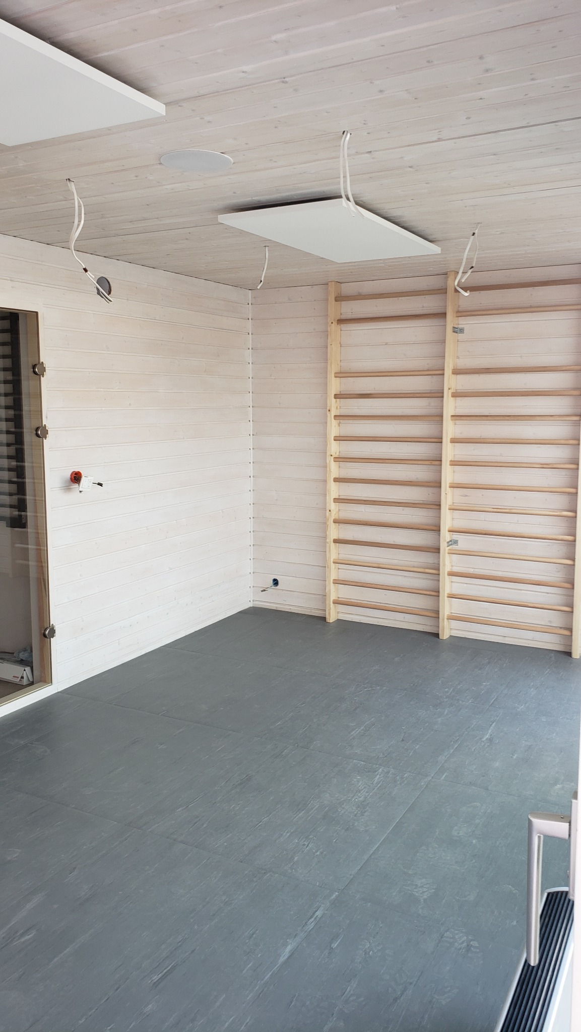 Wnętrze pomieszczenia wykończonego drewnem z drabinkami gimnastycznymi, szarą podłogą i panelami sufitowymi w trakcie montażu oświetlenia.