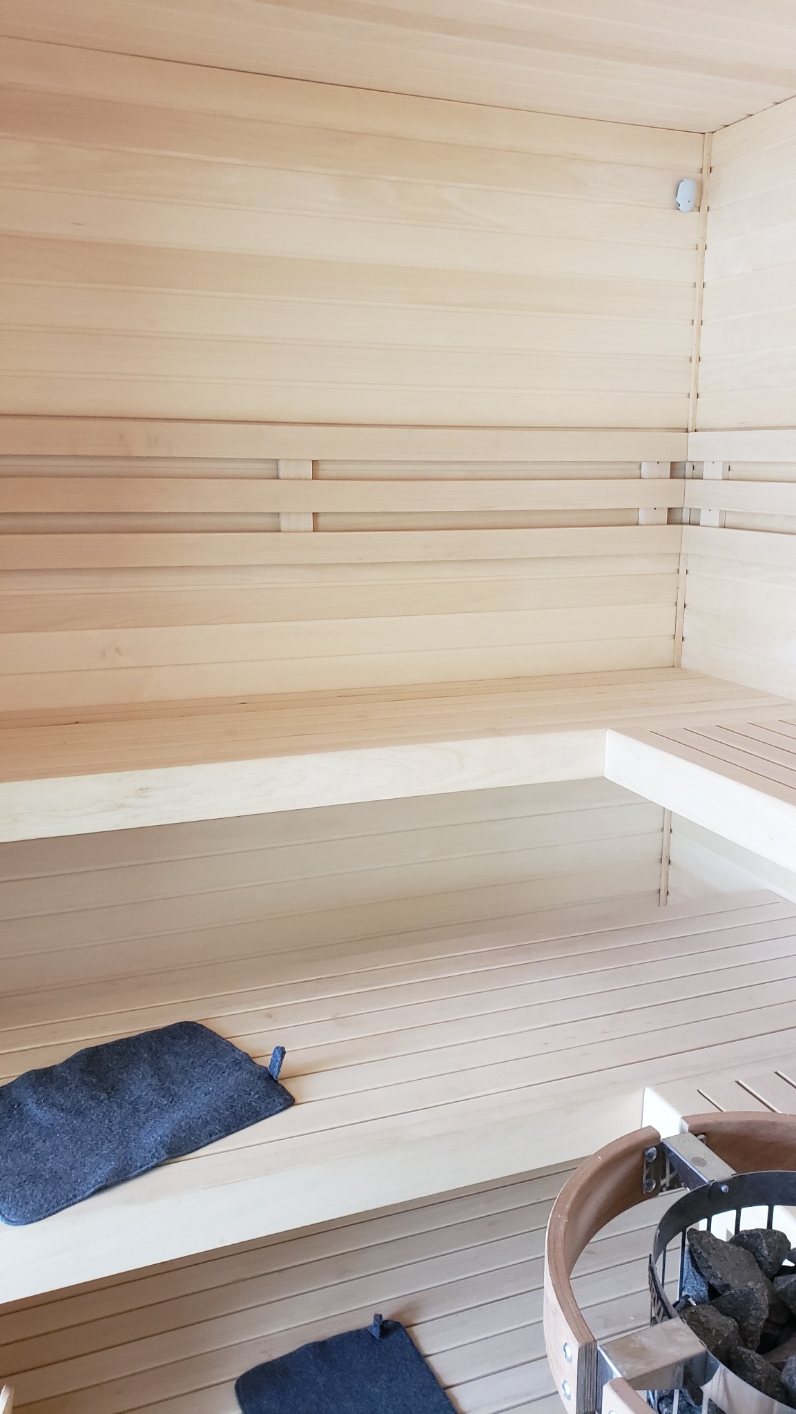 Wnętrze nowej, jasnej sauny z drewnianymi ławkami, dwoma ciemnymi podkładkami filcowymi na siedziskach i piecem z kamieniami w drewnianej obudowie.