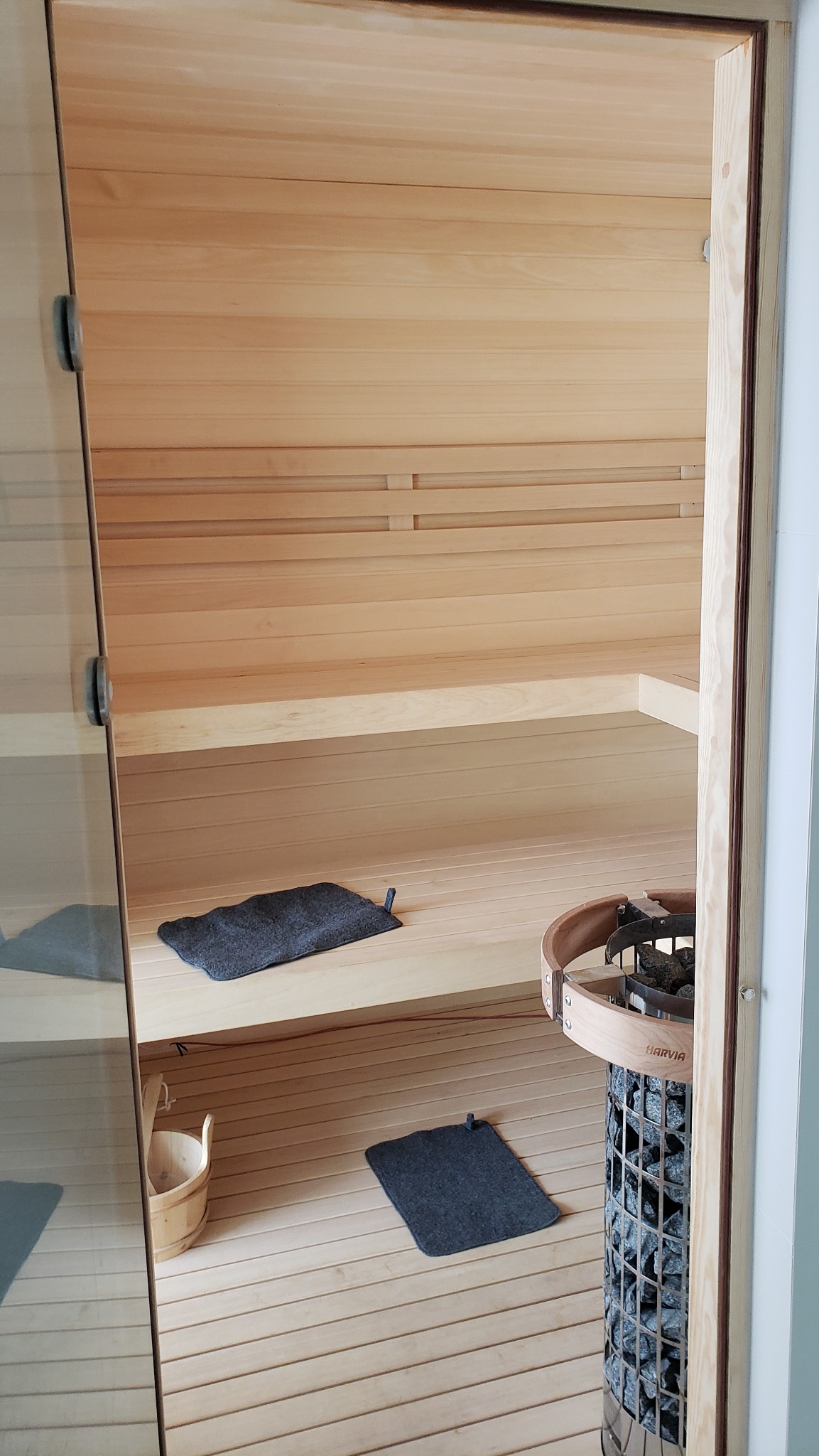 Wnętrze wykończonej sauny z jasnego drewna, z piecem Harvia wypełnionym kamieniami, drewnianym wiadrem i dwoma ciemnymi podkładkami filcowymi na ławkach i podłodze.