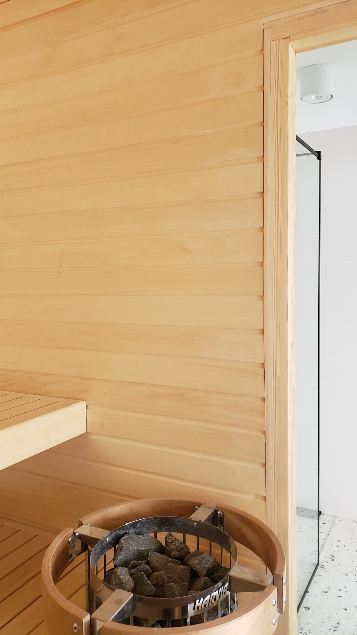 Wnętrze wykończonej sauny z piecem na kamienie marki Harvia, drewniane panele ścienne i ławka, widok na szklane drzwi kabiny prysznicowej w tle.