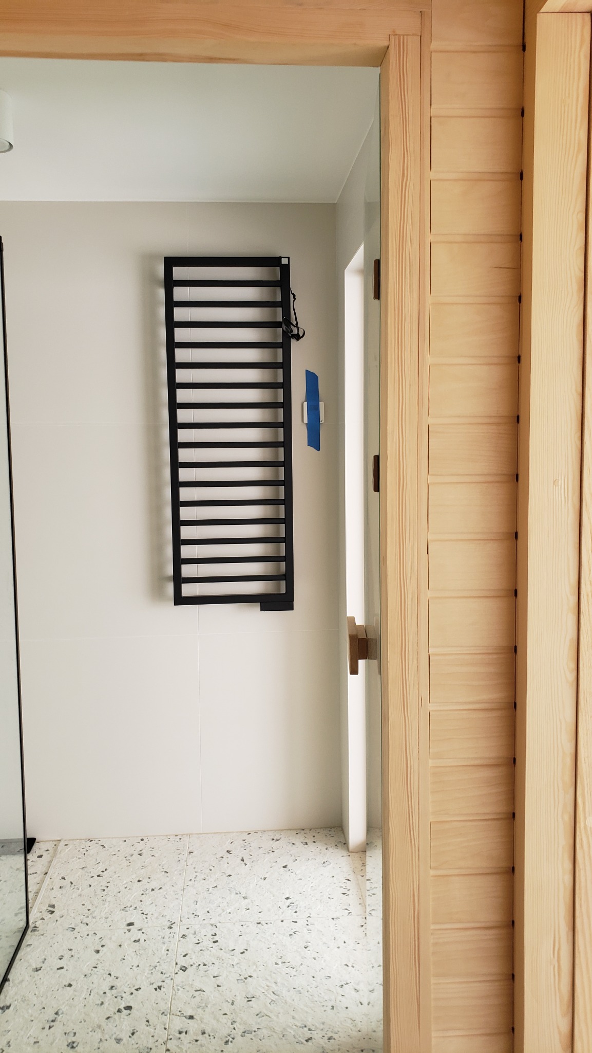 Fragment łazienki z grzejnikiem drabinkowym w kolorze czarnym, zamontowanym na białej ścianie wyłożonej kafelkami. Po prawej stronie widoczna drewniana konstrukcja z jasnego drewna.