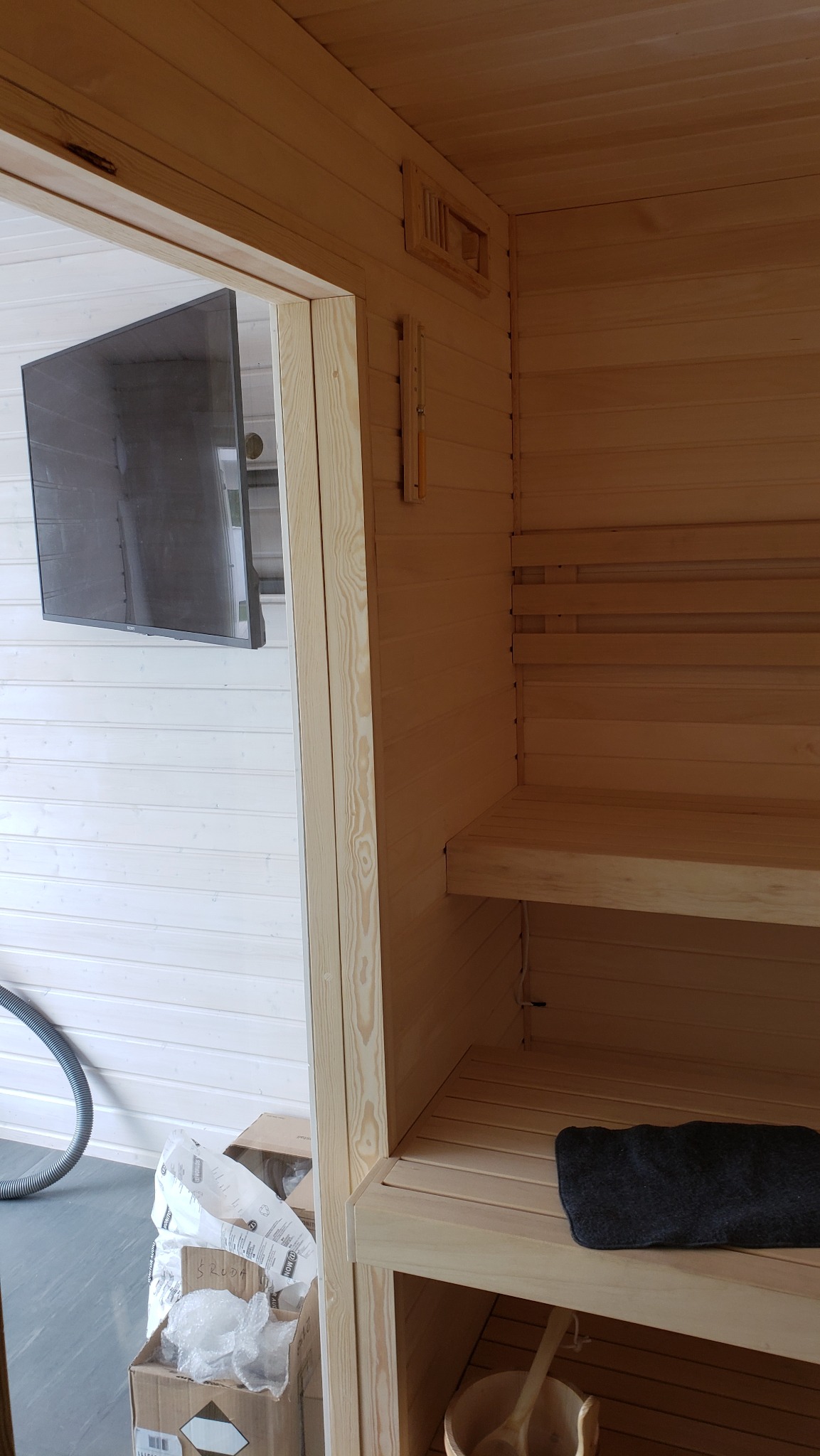 Wnętrze sauny z drewnianymi ławkami i termometrem na ścianie, widoczny telewizor na zewnątrz i kartonowe pudełka z opakowaniami po materiałach budowlanych.