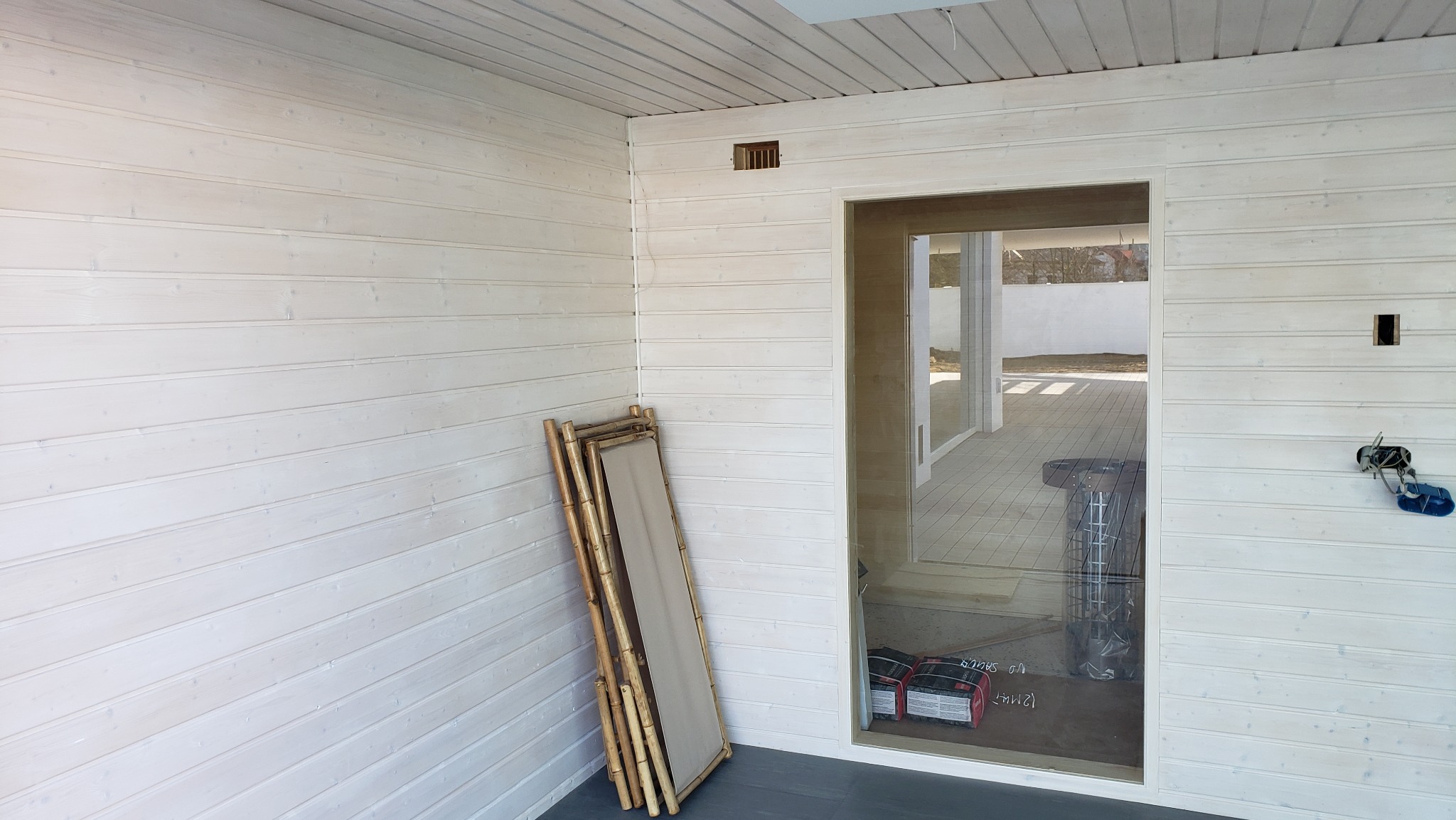Wnętrze wykończone białymi panelami boazeryjnymi, widoczny otwór wentylacyjny, instalacja elektryczna w trakcie montażu oraz złożony leżak bambusowy oparty o ścianę obok przeszklonych drzwi.