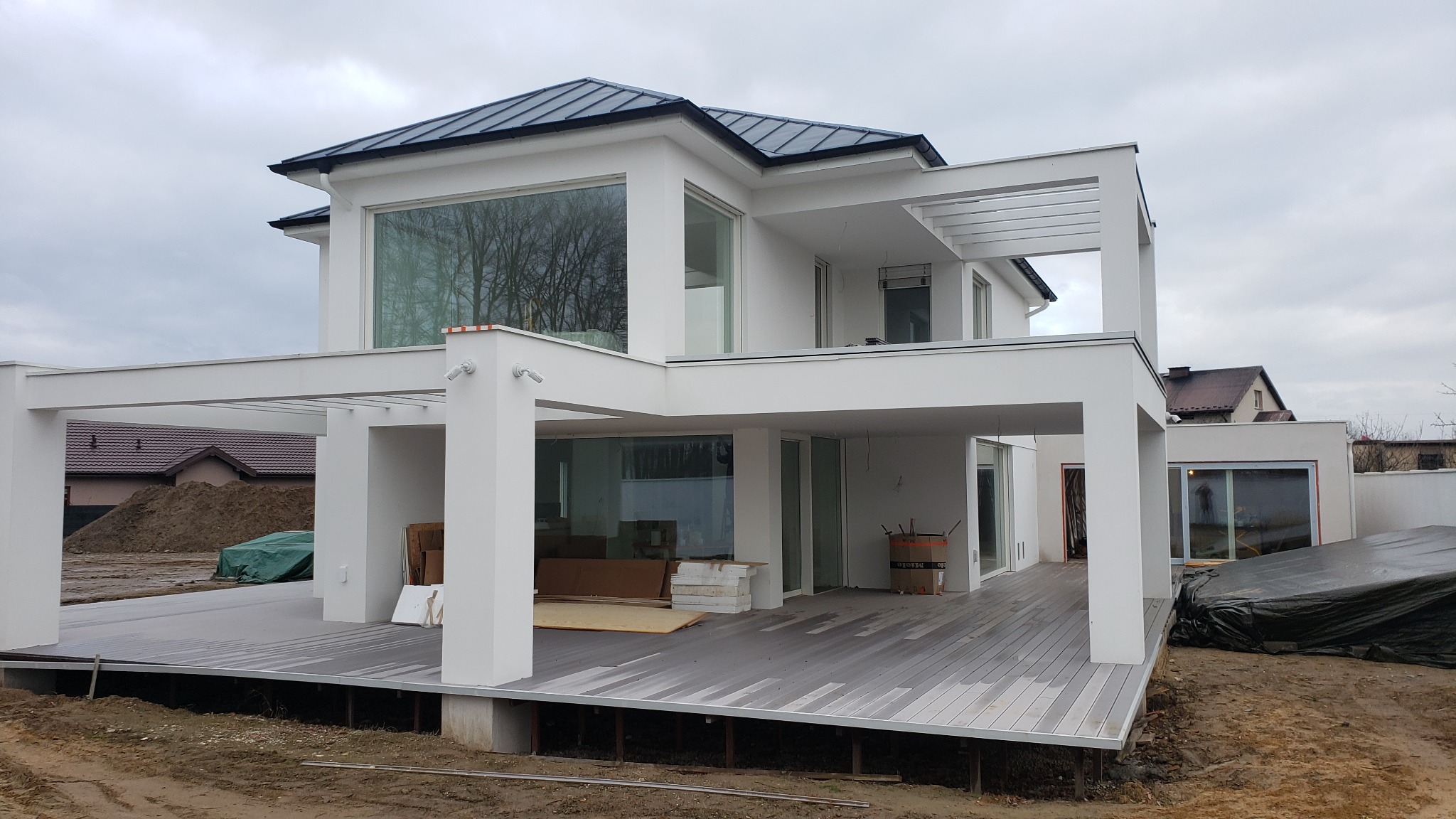 Nowoczesny, dwukondygnacyjny dom z płaskim dachem i dużymi oknami, zadaszony taras z szarych desek kompozytowych, widoczne prace wykończeniowe i materiały budowlane na zewnątrz.