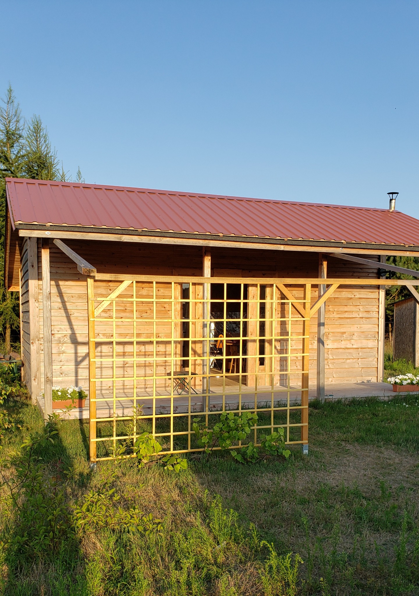 Drewniany budynek z czerwonym dachem i kratownicą z przodu, otoczony zielenią.