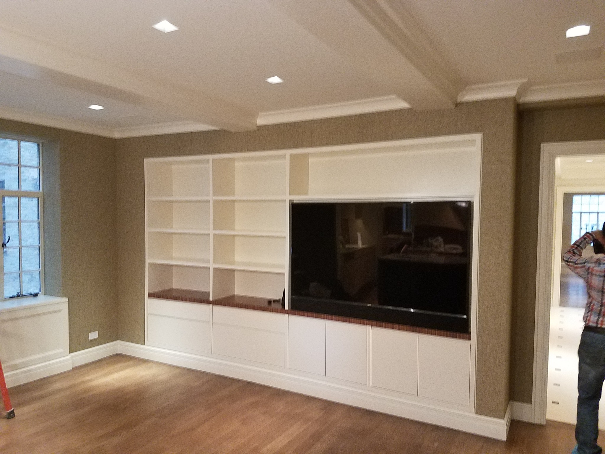 Biała zabudowa meblowa z półkami i szafkami w salonie z telewizorem, drewniana podłoga, dekoracyjne listwy sufitowe, ściany w odcieniach beżu, widoczna część drabiny malarskiej.