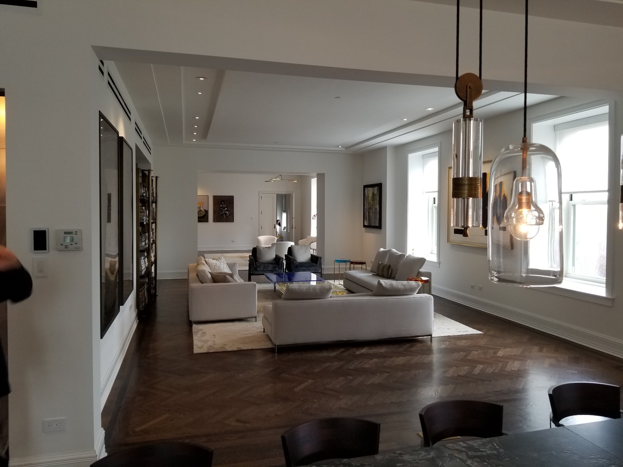 Przestronne wnętrze salonu z parkietem w jodełkę, jasnymi ścianami i nowoczesnym oświetleniem wiszącym, widok na dalszą część pomieszczenia z sofami i fotelami.