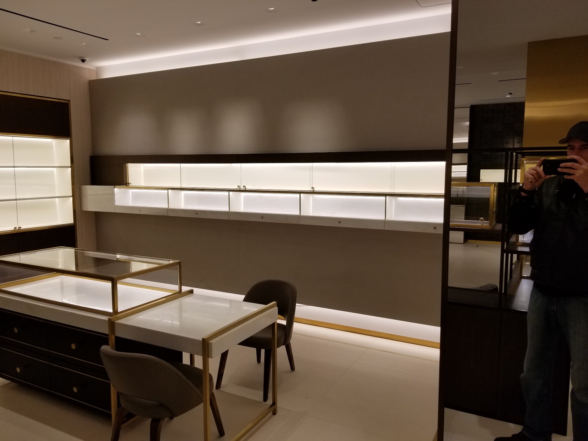 Wnętrze sklepu z oświetlonymi witrynami i meblami w stylu art deco, widoczne krzesła i odbicie osoby robiącej zdjęcie.