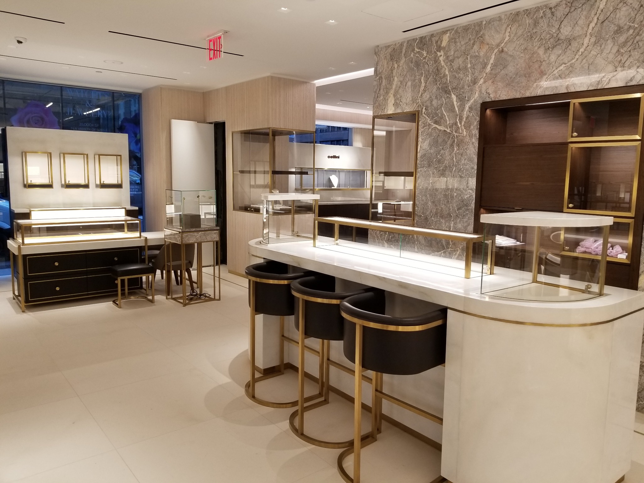 Wnętrze luksusowego sklepu w Zduńskiej Woli z witrynami i regałami na biżuterię, wykonanymi z białego marmuru i złota, oraz czarnymi krzesłami barowymi.