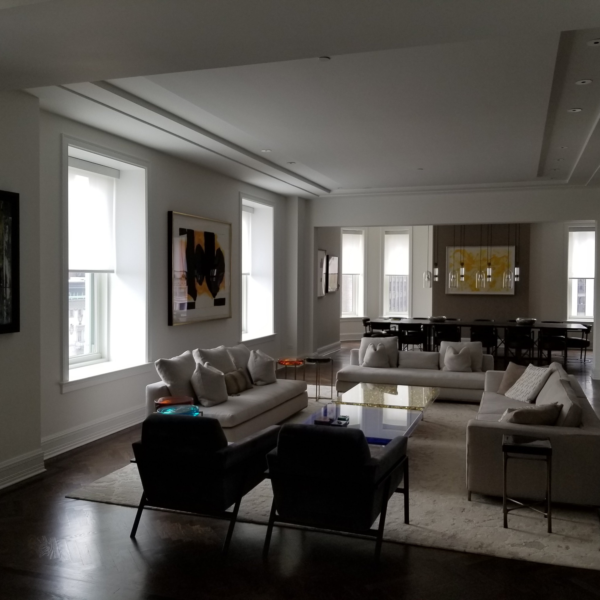 Przestronny salon w odcieniach bieli i szarości, z trzema sofami, ciemnymi fotelami i parkietem w jodełkę. Duże okna z roletami wpuszczają naturalne światło. W tle widoczna jadalnia z długim stołem...
