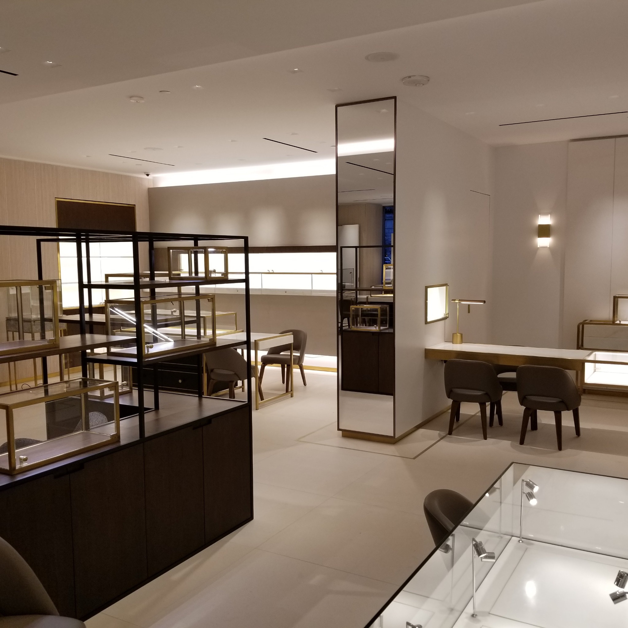 Wnętrze sklepu z ekspozycją mebli i oświetleniem, prezentujące metalowe regały z przeszklonymi gablotami oraz biurko z krzesłami w neutralnej kolorystyce.